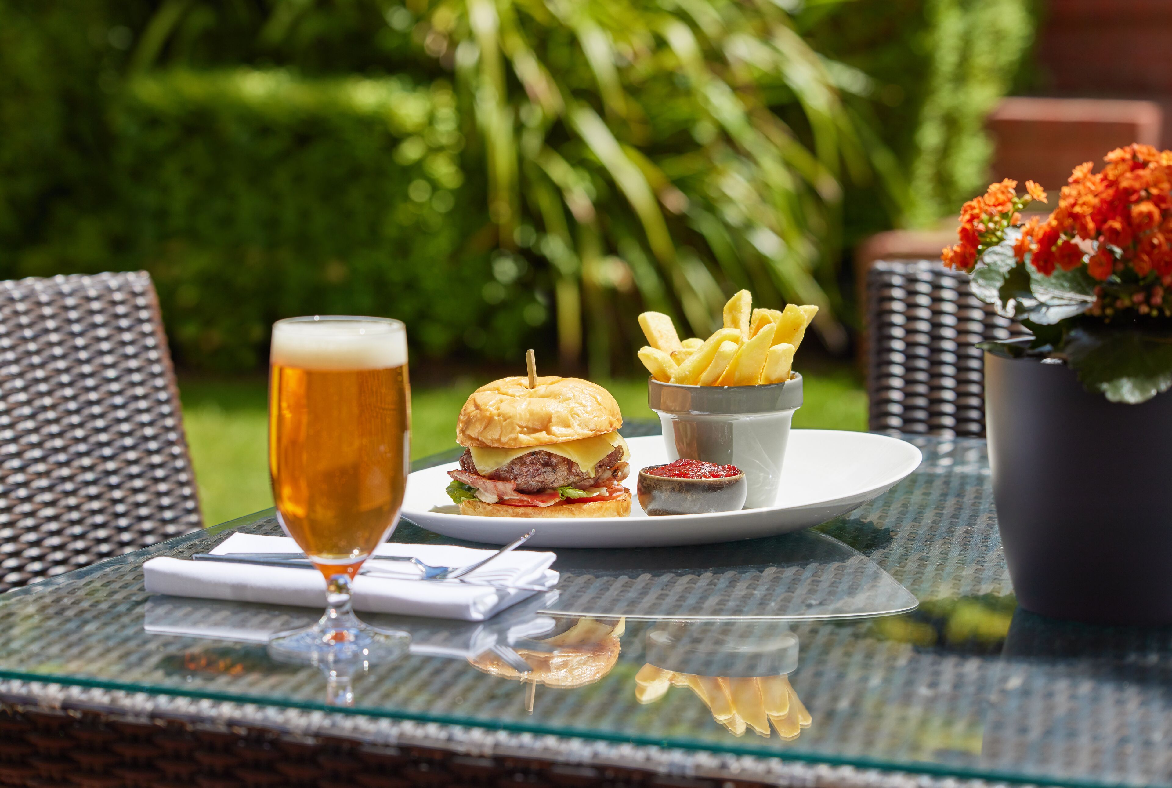 A glass of beer, a burger, and French fries on a glass table outdoors.