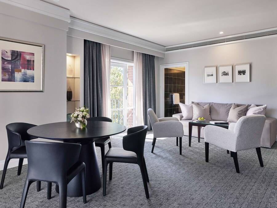 Une chambre d’hôtel moderne avec une table à manger ronde, des chaises noires, un canapé gris et une moquette à motifs.
