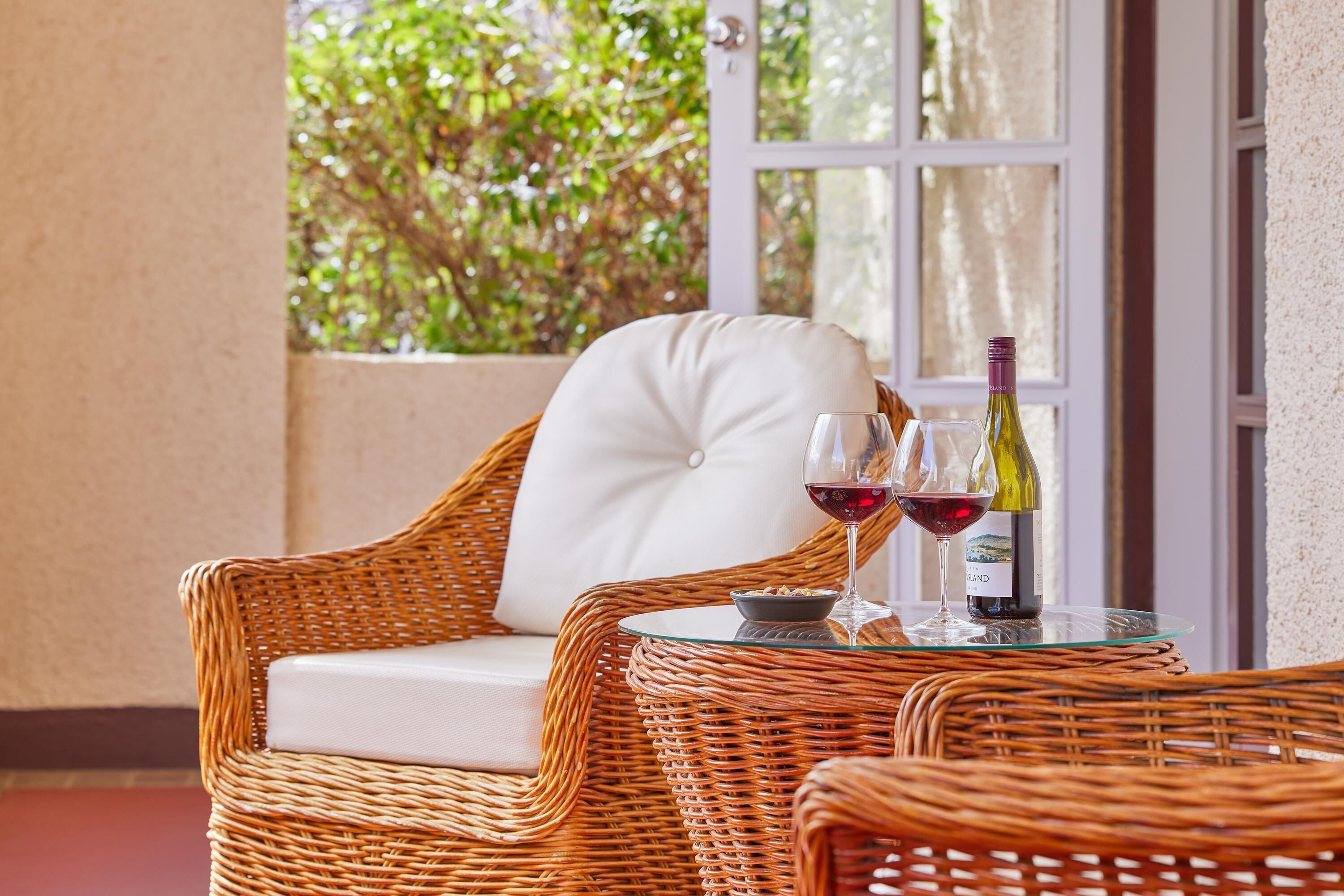 A wicker chair with a white cushion, a glass table with two wine glasses and bottles, and a view of greenery through a window.