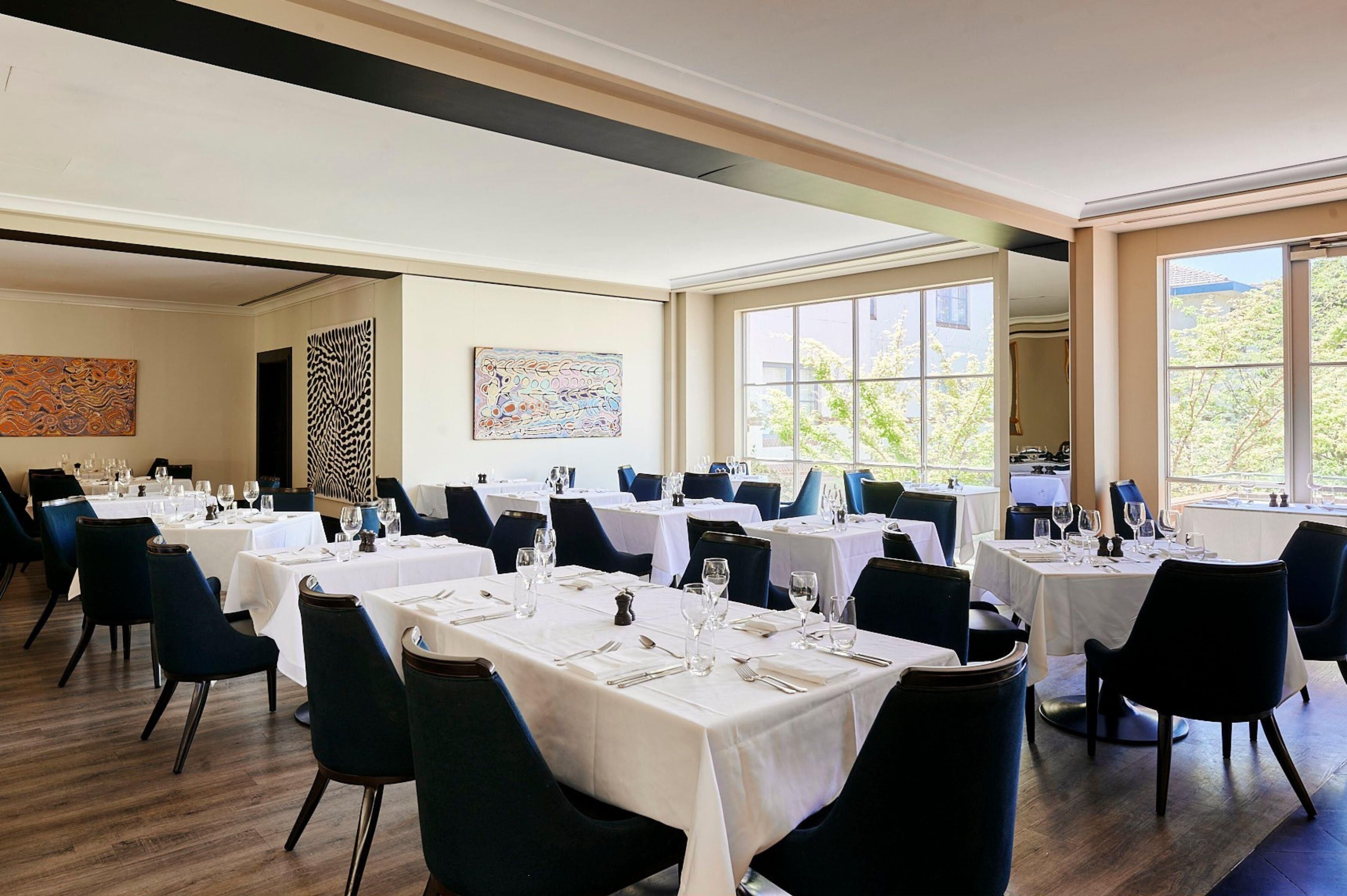 A dining area with multiple tables set for service, large windows with a view of greenery, and artwork on the walls.