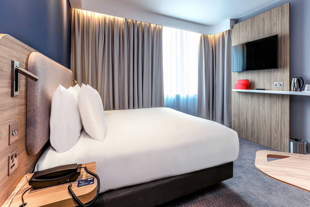 A modern hotel room with a large bed, a mounted TV, a bedside table with a phone, and a small desk area.