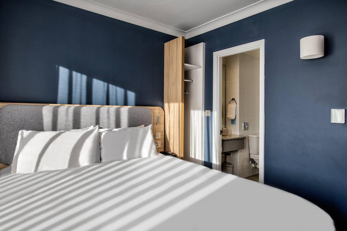 A modern hotel room with a large bed, a wooden headboard, a built-in wardrobe, and a bathroom visible through an open door.