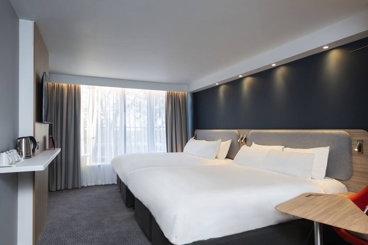 A modern hotel room with a large bed, a bedside table, a desk, and a view of the outdoors through the window.