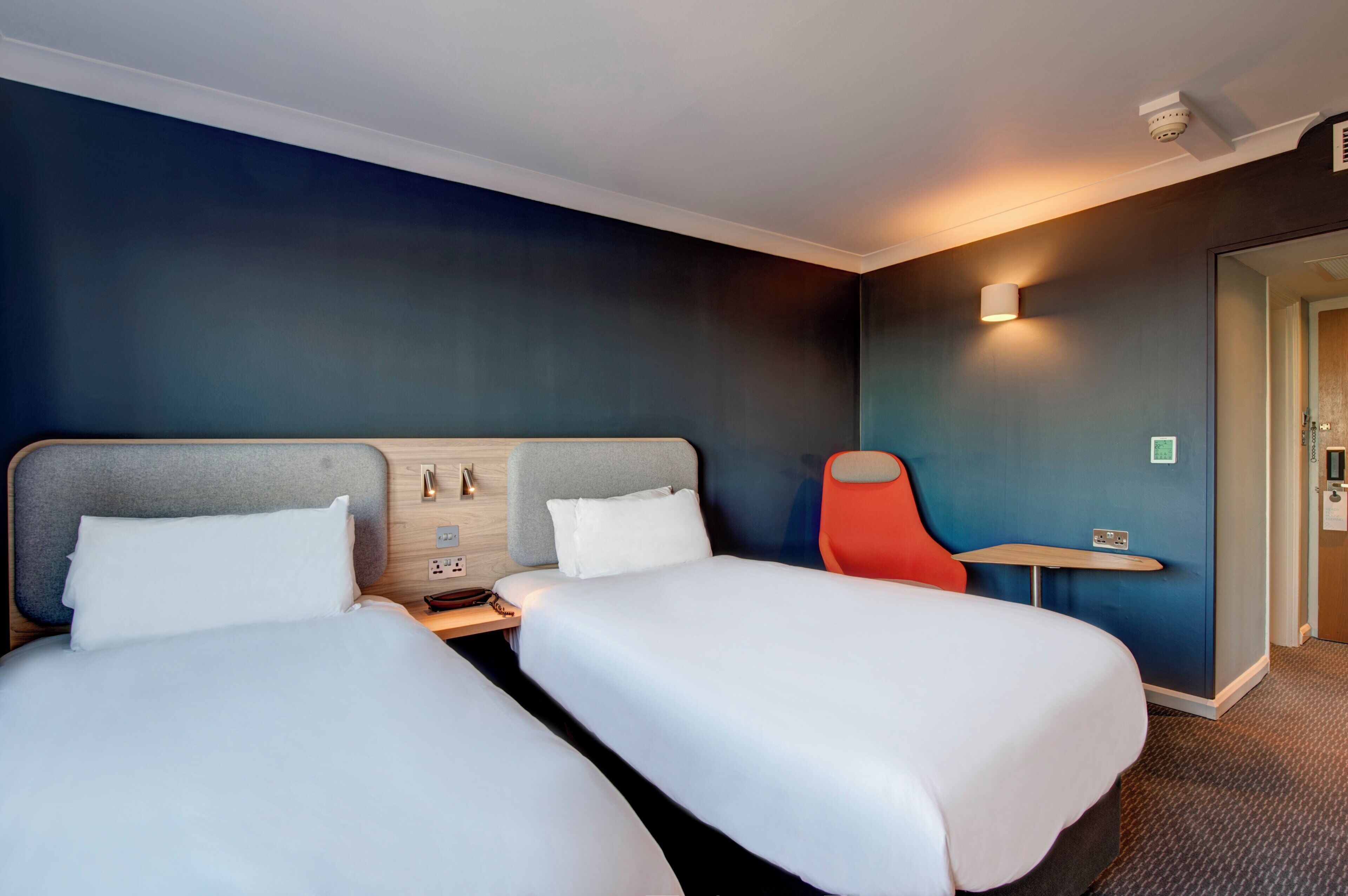 A hotel room with two beds, a small table, a chair, and a wall-mounted light fixture.