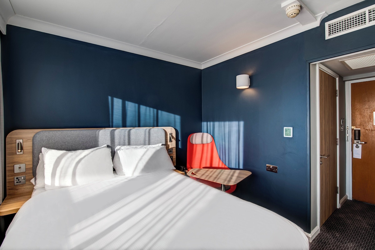 Hotellihuone, jossa on suuri sänky, puinen yöpöytä, punainen tuoli, seinään kiinnitetty lamppu ja ovi, jossa on numeronäppäimellä avattava lukko.