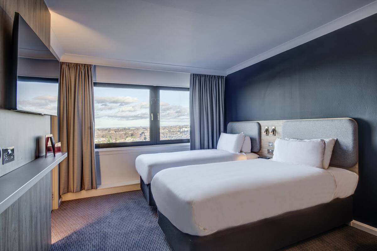 A hotel room with two beds, a large window with curtains, a desk with a lamp, and a view of the city.