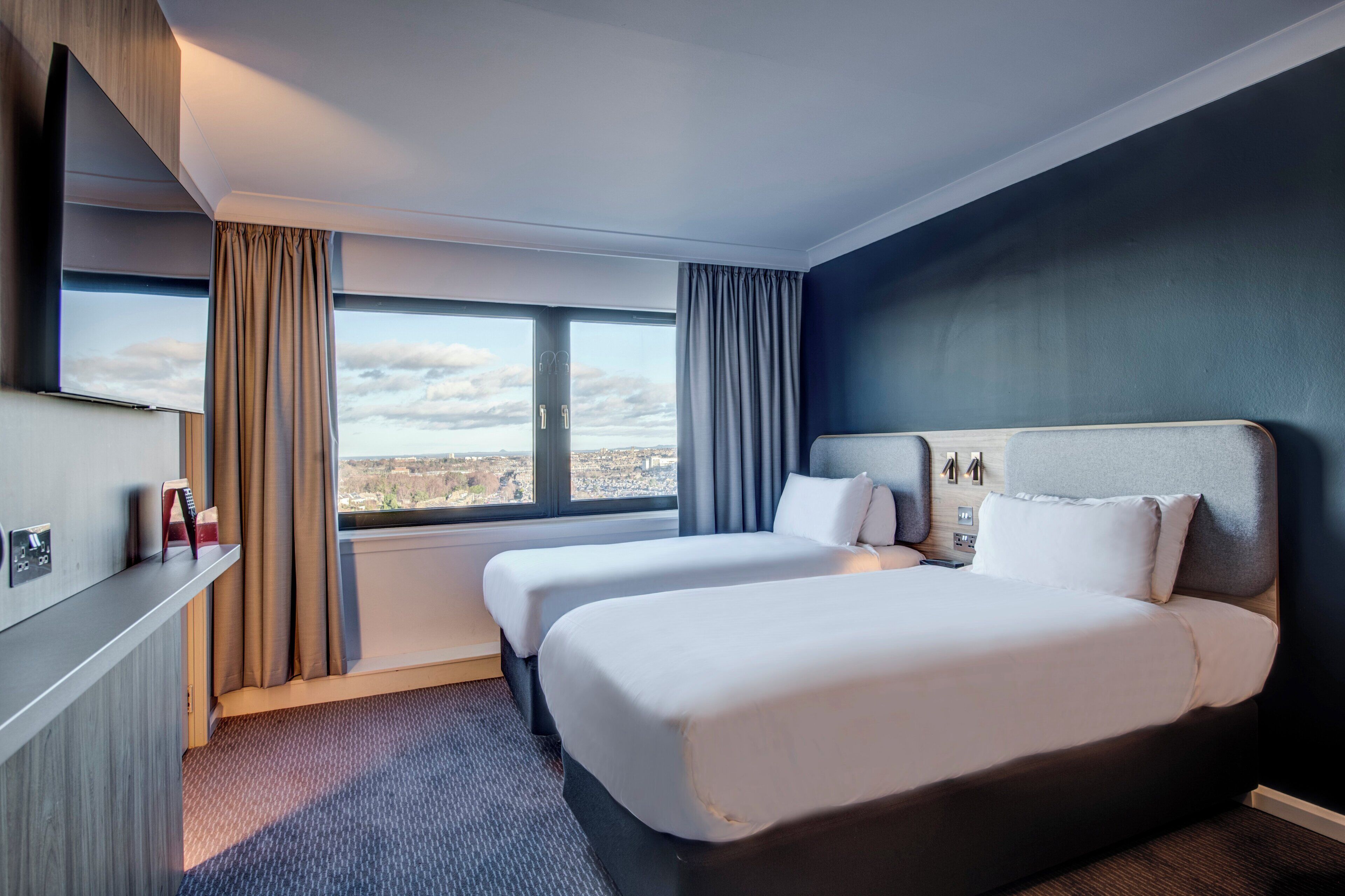 Habitación de hotel con dos camas, un ventanal con cortinas, un televisor de pantalla plana y vistas a la ciudad.