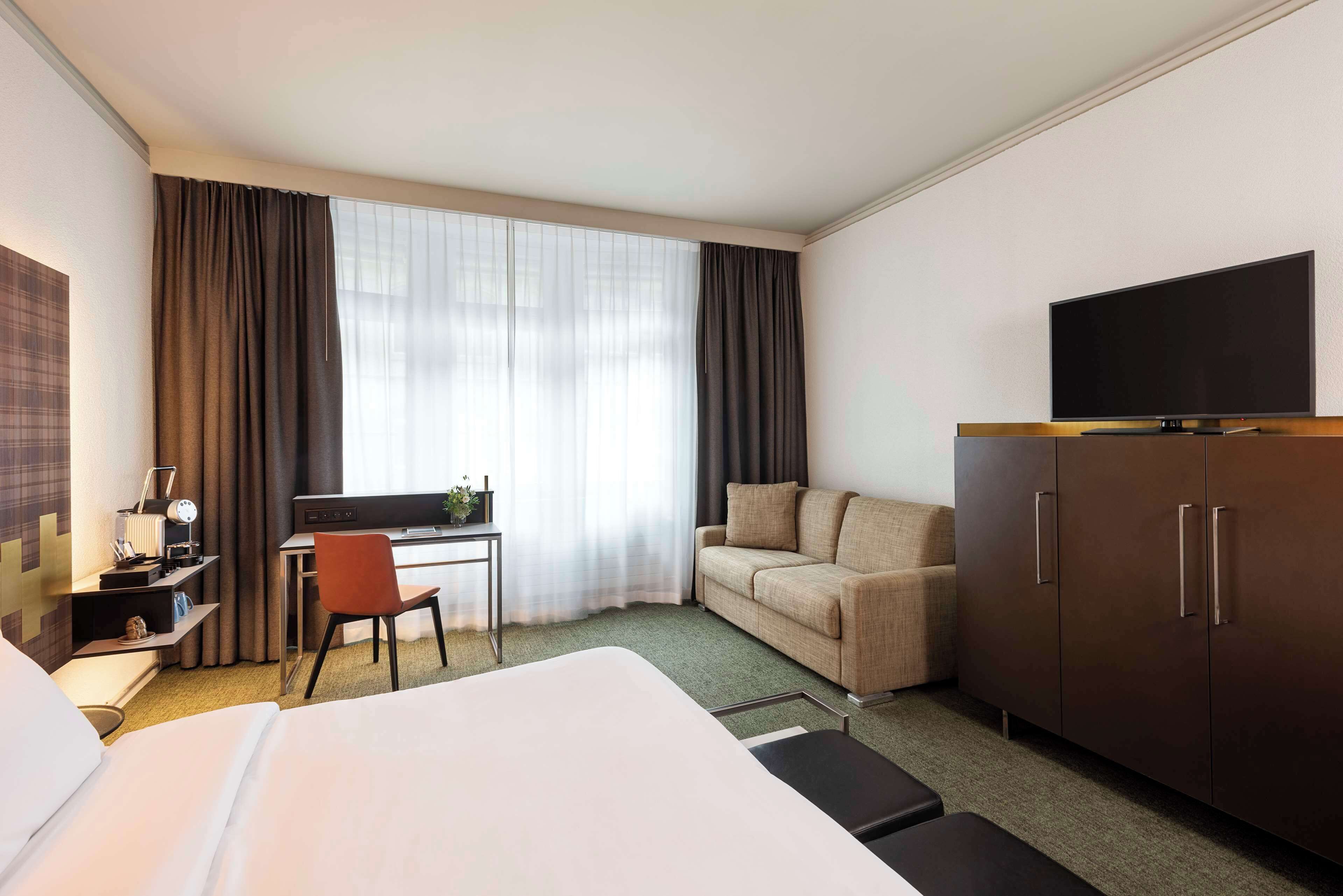 A hotel room with a bed, a desk with a chair, a sofa, a TV on a cabinet, and a large window with curtains.