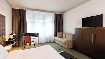 A hotel room with a bed, a desk with a chair, a sofa, a TV on a cabinet, and a large window with curtains.