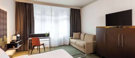A hotel room with a bed, a desk with a chair, a sofa, a TV on a cabinet, and a large window with curtains.