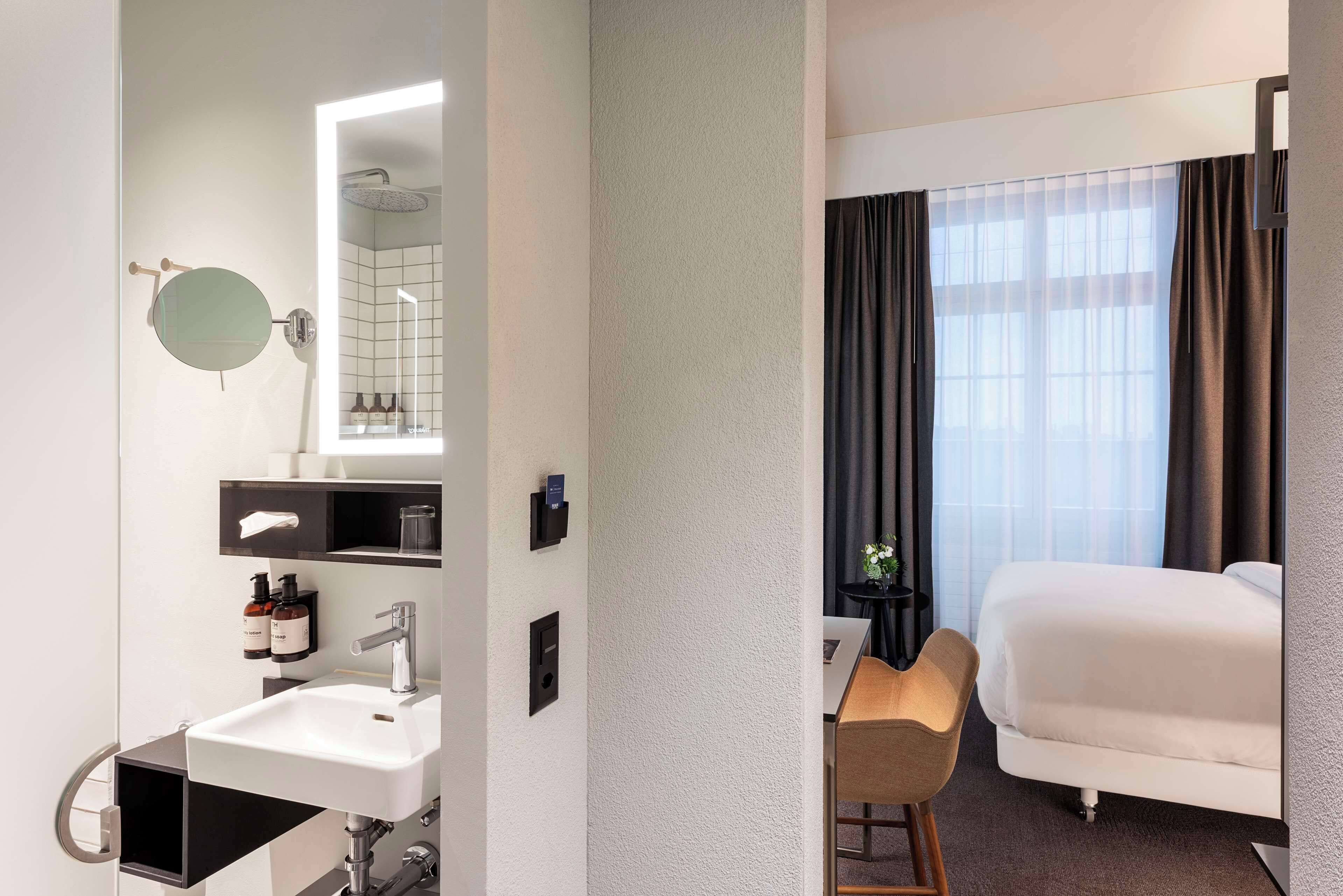 A hotel room with a bed, a desk, a chair, a sink, a mirror, and a window with curtains.