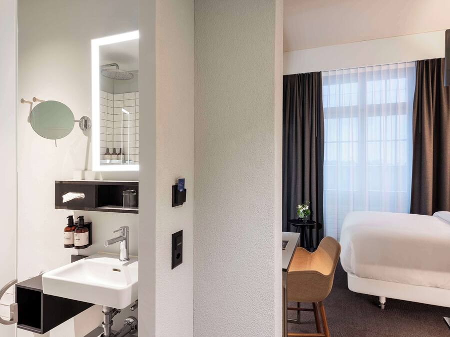 Habitación de hotel con cama, escritorio, silla, lavamanos, espejo y ventana con cortinas.