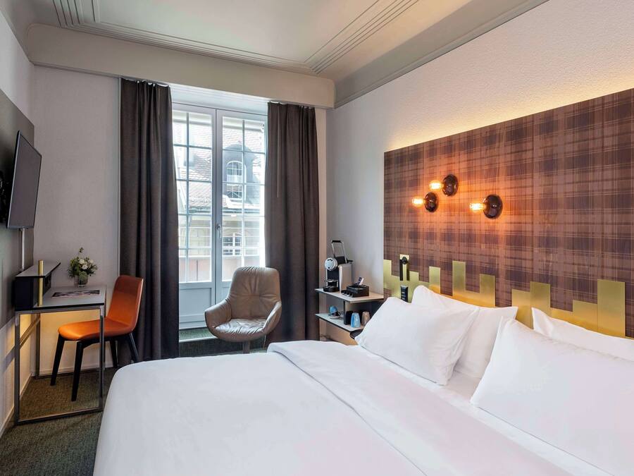 Ett hotellrum med en stor säng, ett skrivbord med en stol, en tv och ett fönster med gardiner.