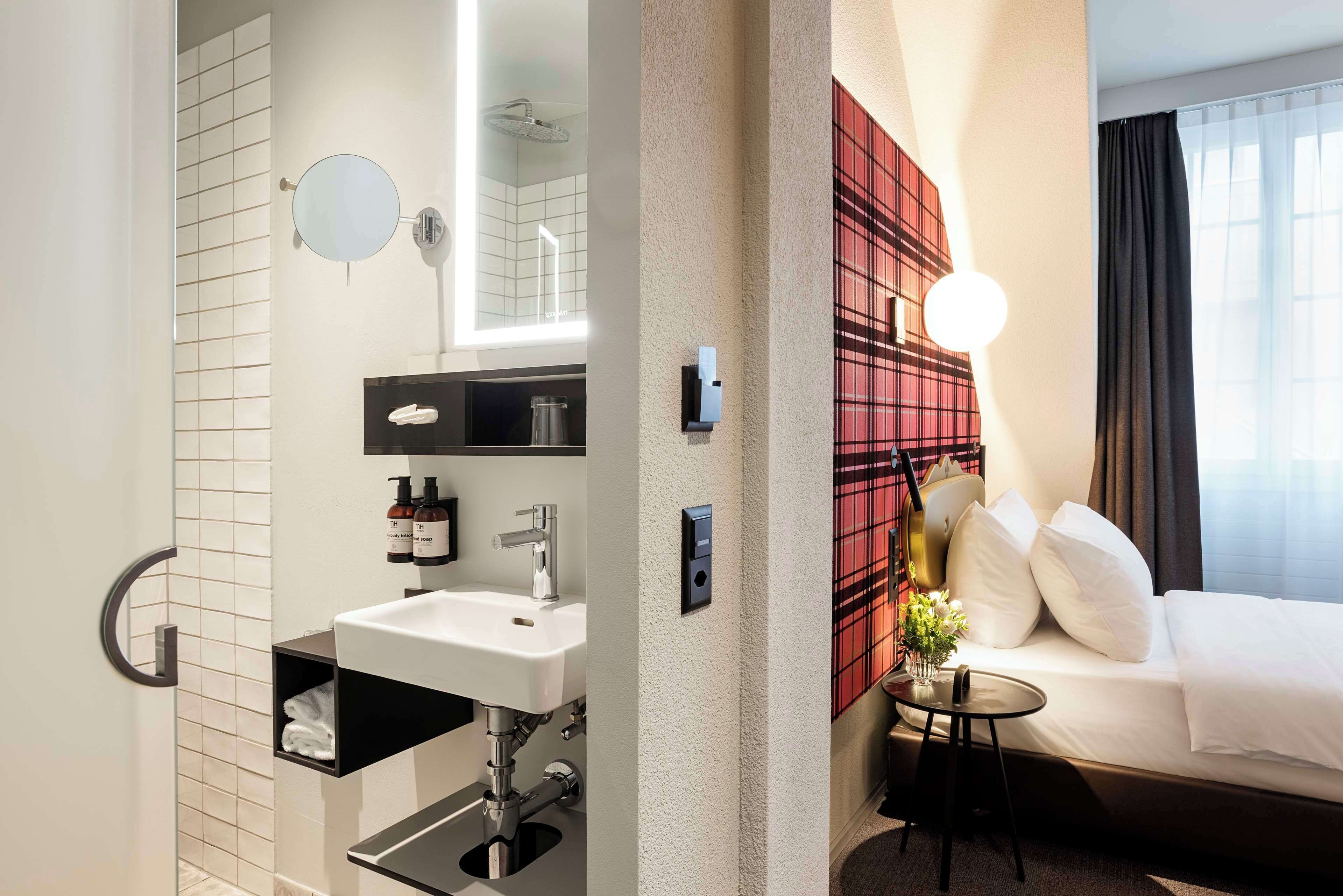 A modern bathroom with a sink, mirror, and towel rack. The room features a bed with white bedding and a small bedside table with a lamp.