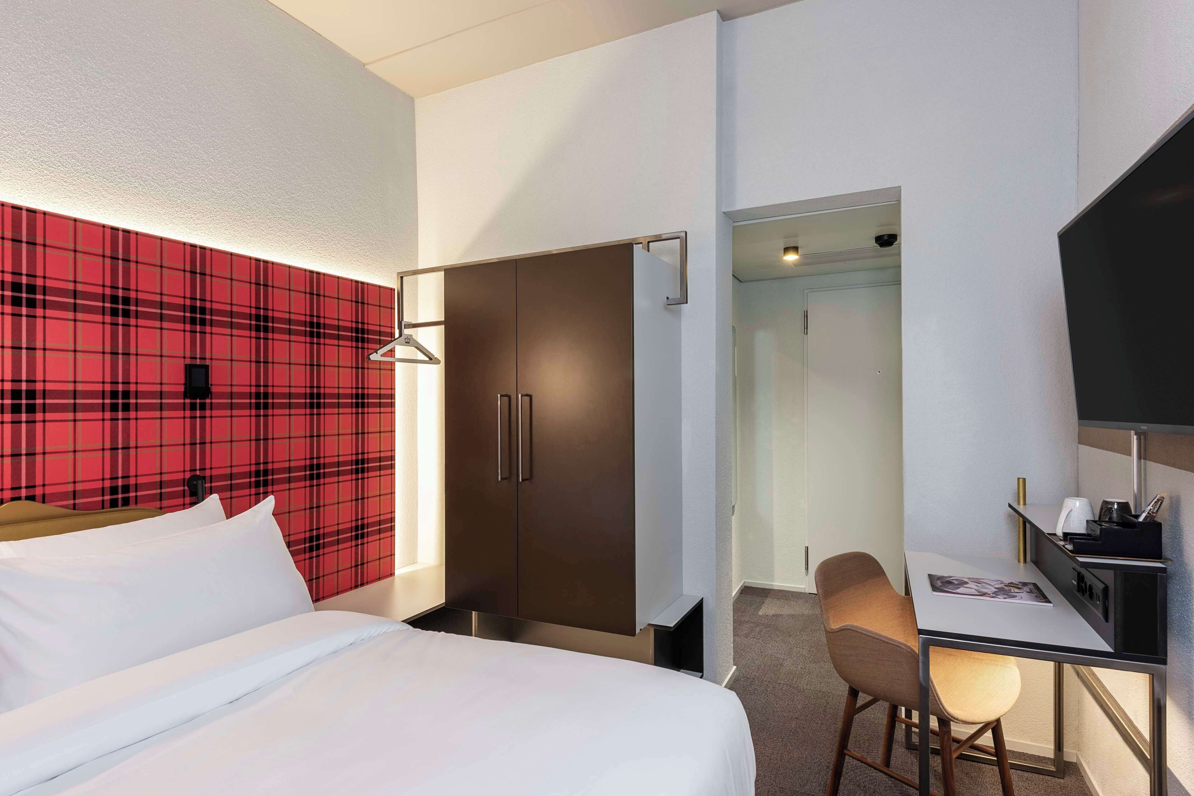 Una habitación de hotel con cama, escritorio y silla, armario y una pared con estampado escocés rojo.
