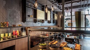 A modern restaurant with a variety of food and drink options, including bowls of fruit, bread, and salad. - NH Bern The Bristol (Bern)
