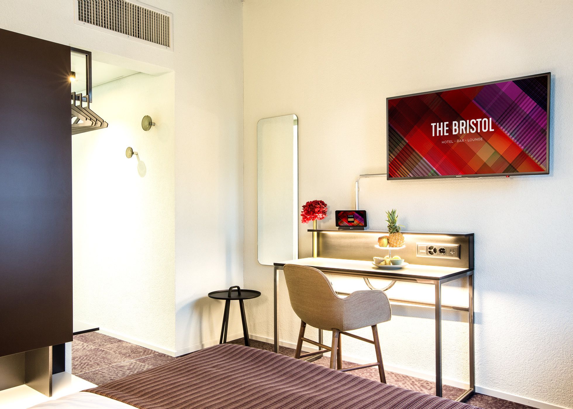A modern hotel room with a desk, chair, and a large mirror. There's a flat-screen TV on the wall displaying "THE BRISTOL HOTEL BAR LOUNGE".