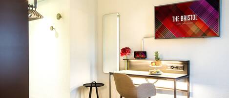 A modern hotel room with a desk, chair, and a large mirror. There's a flat-screen TV on the wall displaying "THE BRISTOL HOTEL BAR LOUNGE".