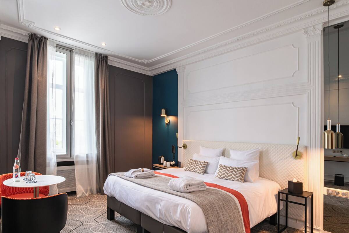 Una habitación de hotel con una cama grande, una mesita de noche, una pequeña mesa redonda con una botella y vasos, y una ventana con cortinas.