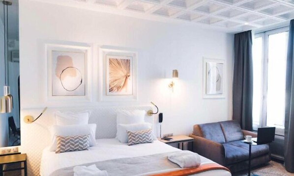 Una habitación de hotel moderna con una cama grande, un sofá y un escritorio. Hay obras de arte enmarcadas en la pared y una ventana con cortinas.
