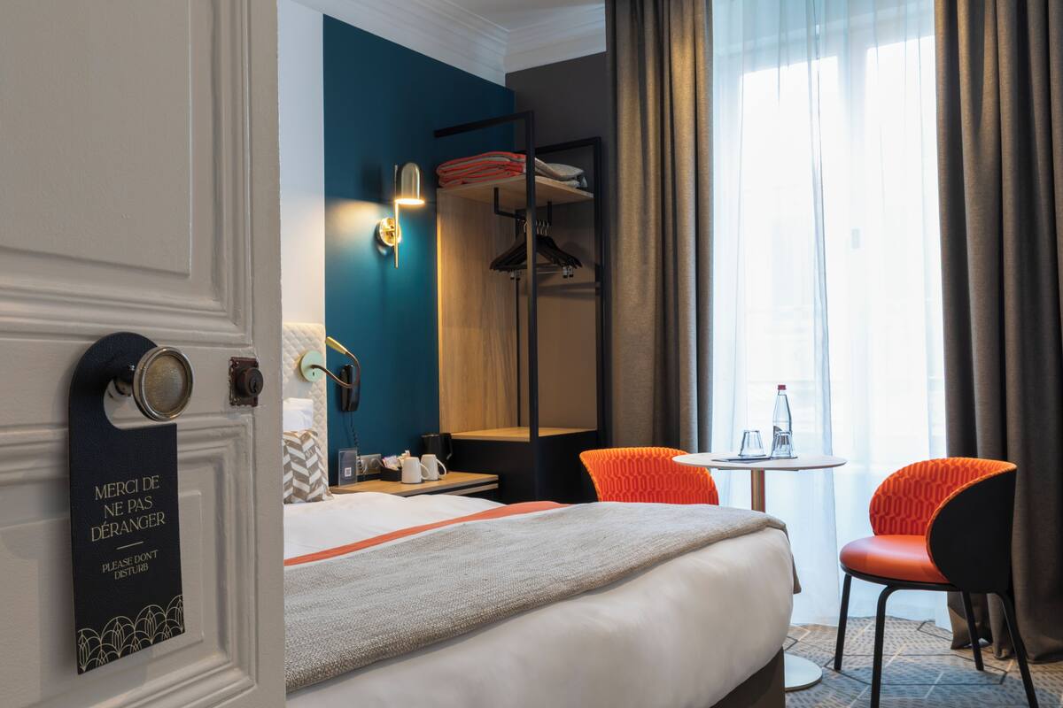 Habitación de hotel con cama, escritorio, silla, mesa, espejo y ventana con cortinas.