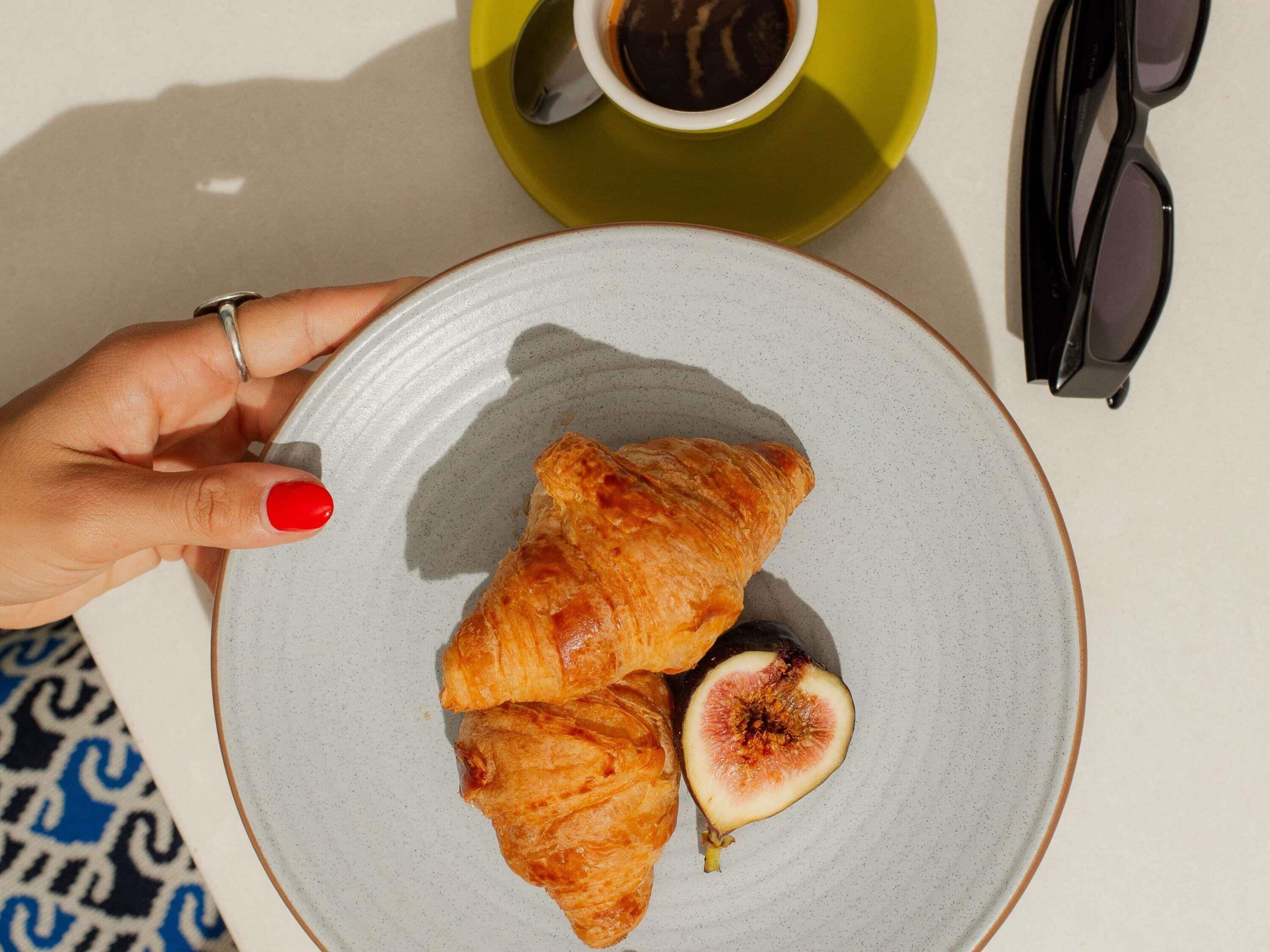 A hand holding a plate with croissants and a fig, a cup of coffee, and sunglasses on a table.