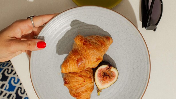 Una mano sosteniendo un plato con croissants y una higo, una taza de café y gafas de sol sobre una mesa.