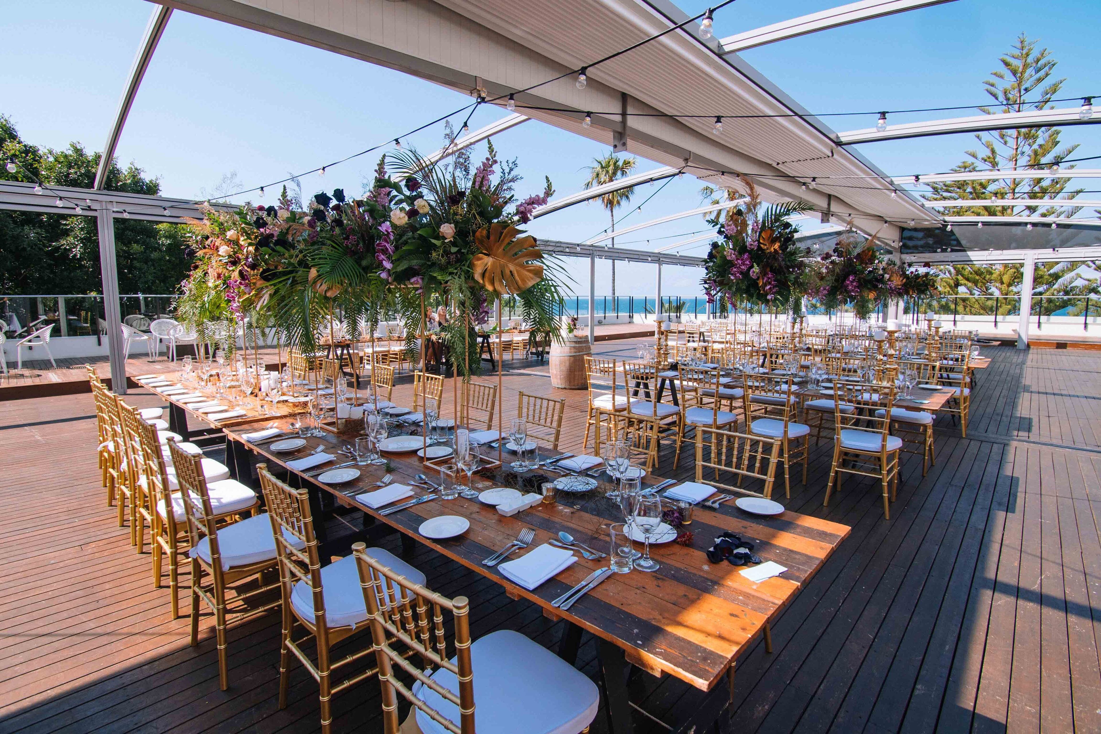 Una larga mesa de madera dispuesta para un evento al aire libre elegante, con sillas doradas, rodeada de exuberantes arreglos florales y una vista despejada del océano.