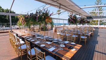 A long wooden table set for a formal outdoor event with gold chairs, surrounded by lush floral arrangements and a clear view of the ocean.