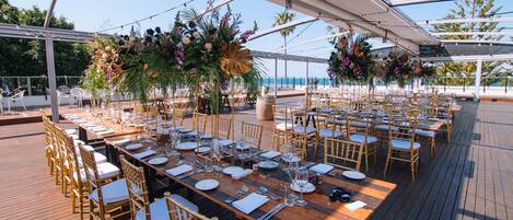 A long wooden table set for a formal outdoor event with gold chairs, surrounded by lush floral arrangements and a clear view of the ocean.