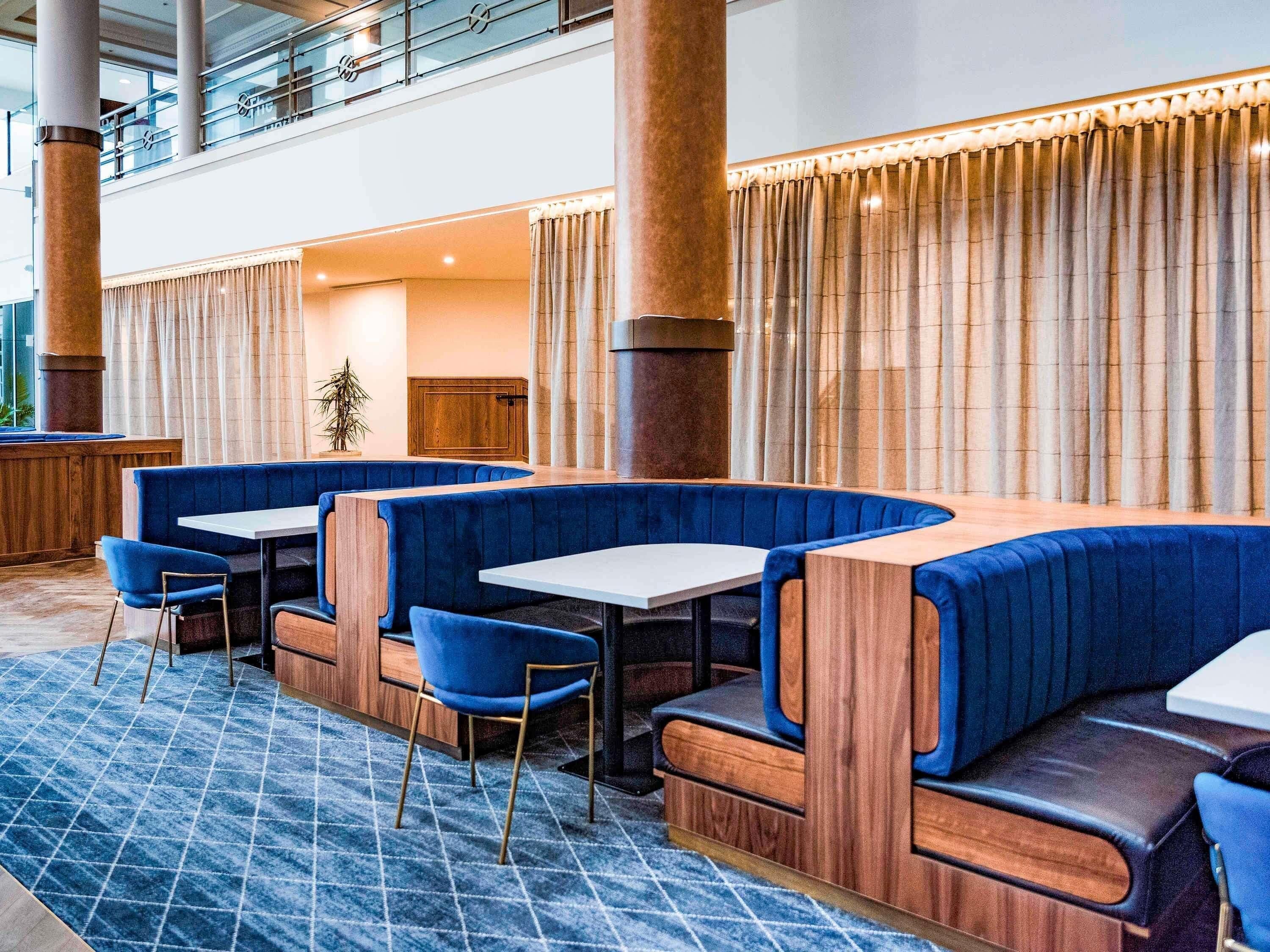 A modern hotel lobby with a seating area featuring blue upholstered booths and tables, large wooden pillars, and a patterned carpet.