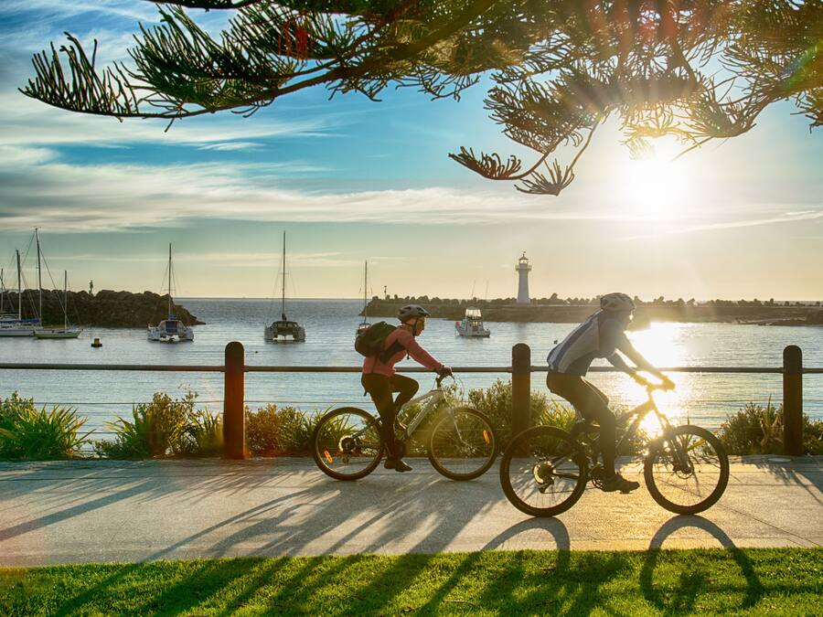 Two cyclists on a paved path by a body of water with boats and a lighthouse in the background.