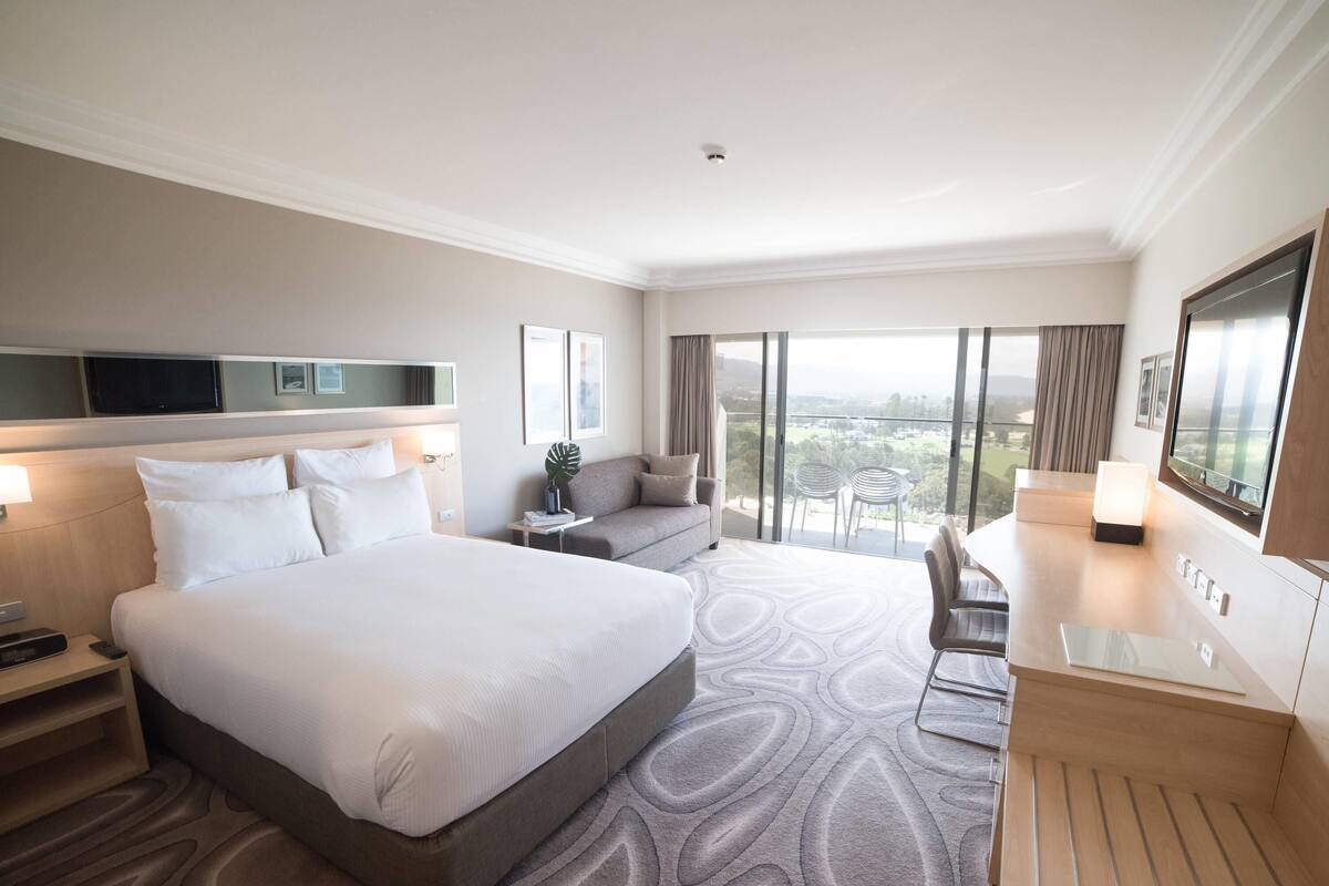 Habitación de hotel con una cama grande, un escritorio, una silla, un televisor y un balcón con vistas.