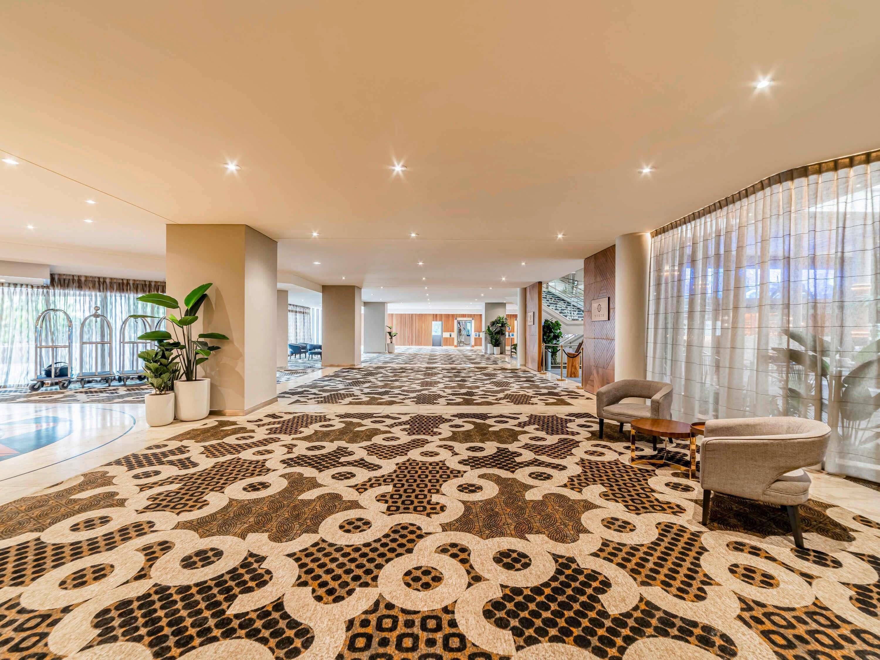Um amplo hall de hotel com carpete estampado, plantas em vasos e uma área de estar.