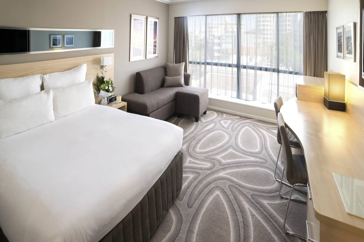 Habitación de hotel con una cama grande, un sofá, un escritorio y vistas a la ciudad a través de amplias ventanas.