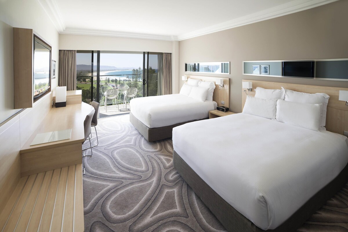 A hotel room with two beds, a desk, a chair, a TV, and a balcony with a view.