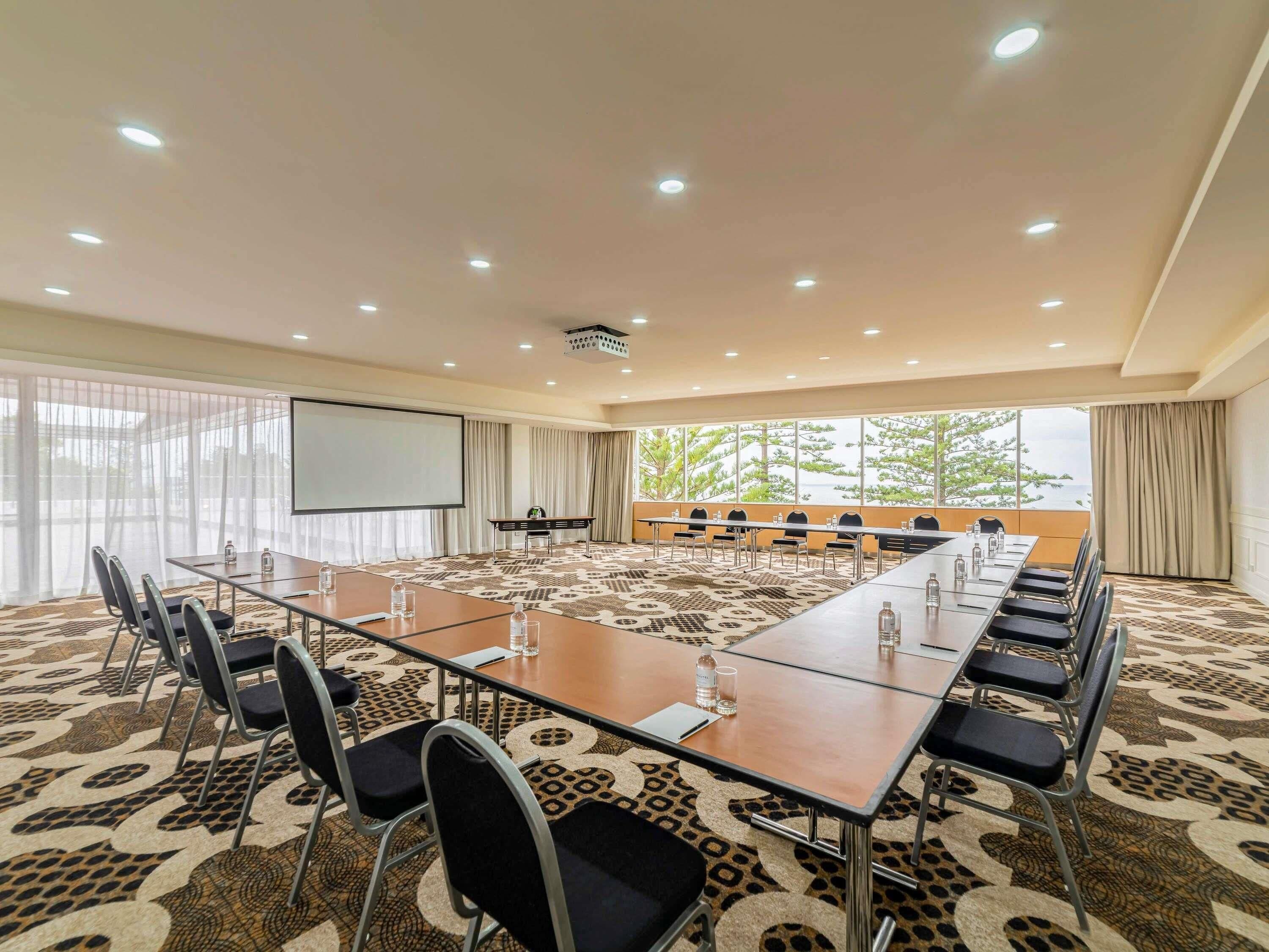 A conference room with a long table, chairs, a projector screen, and a large window with a view.