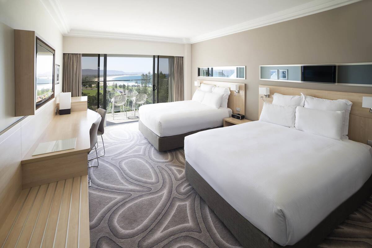 Habitación de hotel con dos camas, un escritorio y un amplio ventanal con vistas al exterior.