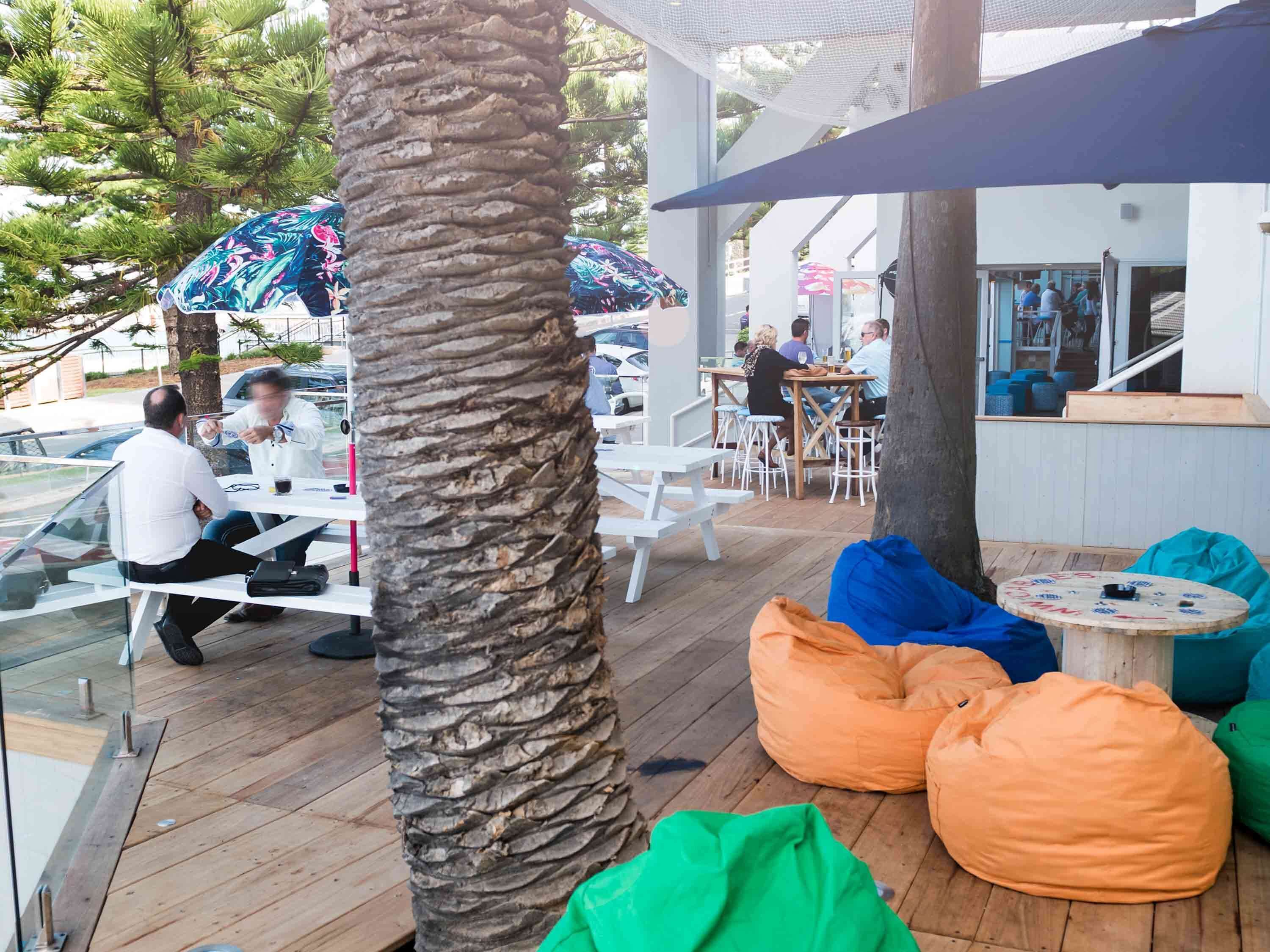 An outdoor seating area with wooden tables and chairs, surrounded by palm trees and colorful umbrellas.