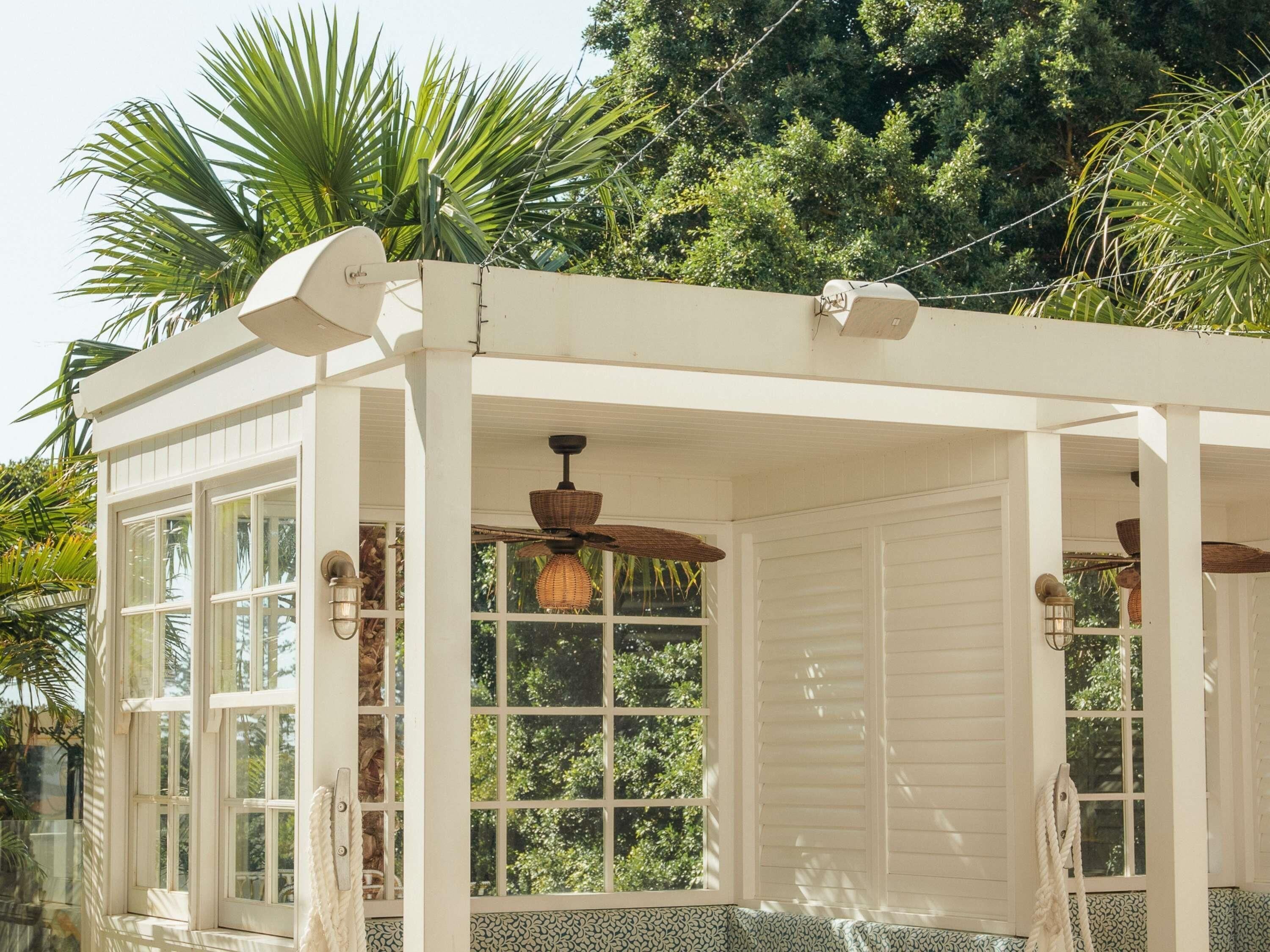 Een witte pergola met grote ramen, een plafondventilator en uitzicht op weelderige begroeiing.