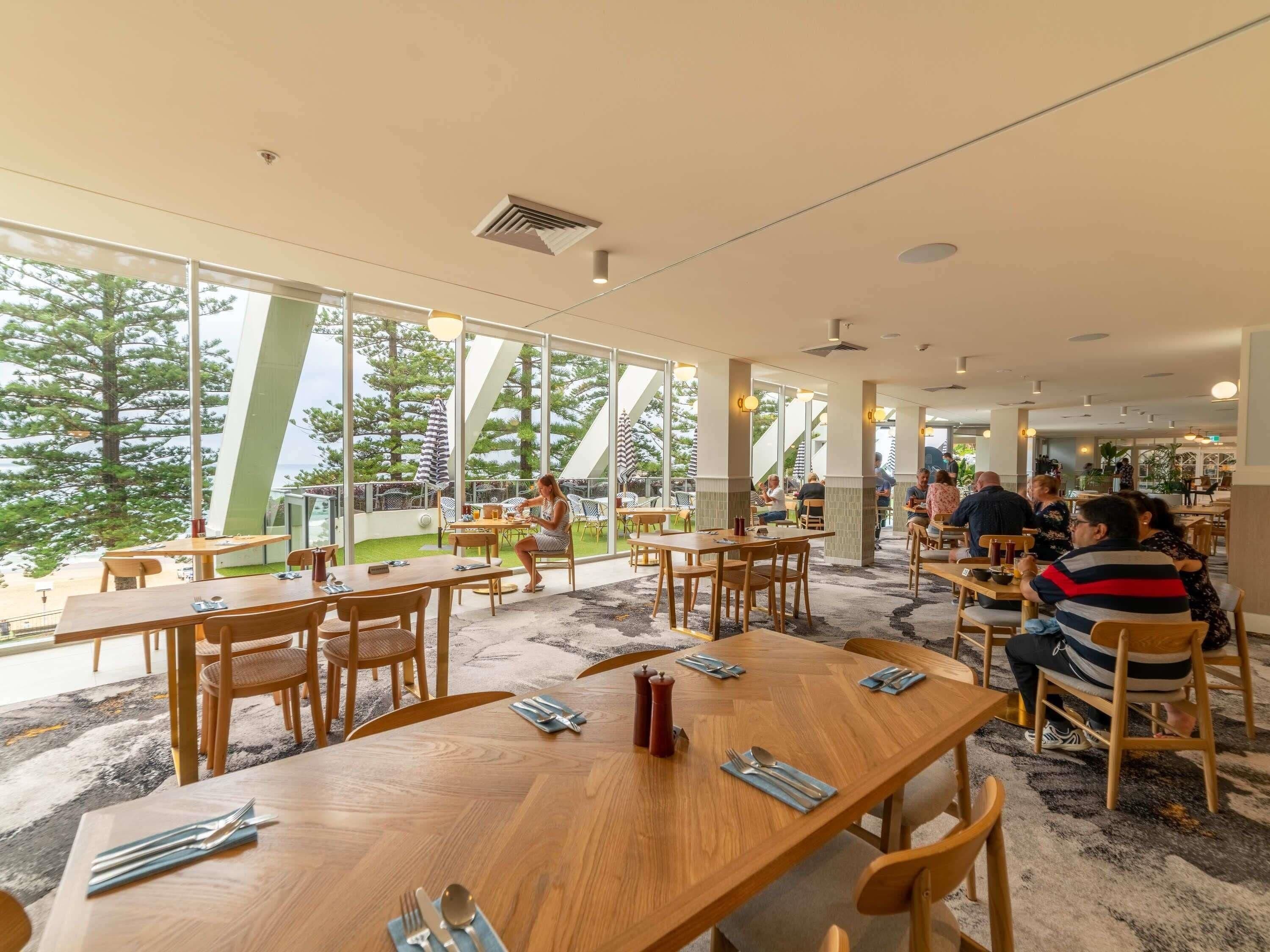 A restaurant with wooden tables and chairs, large windows with a view of trees, and a modern interior design.