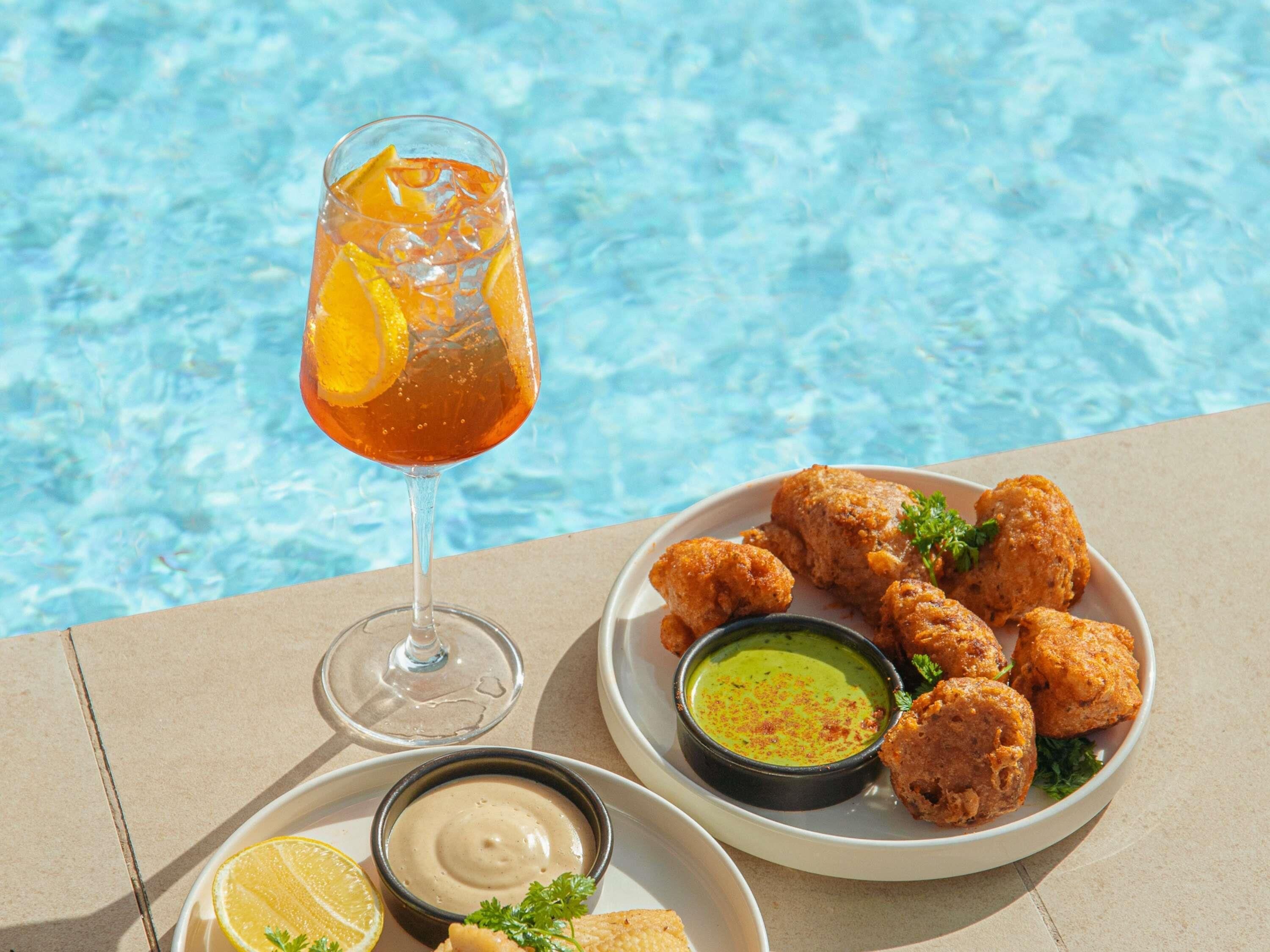 A glass of orange drink with lemon slices, a plate of fried items, and two dipping sauces by a poolside.
