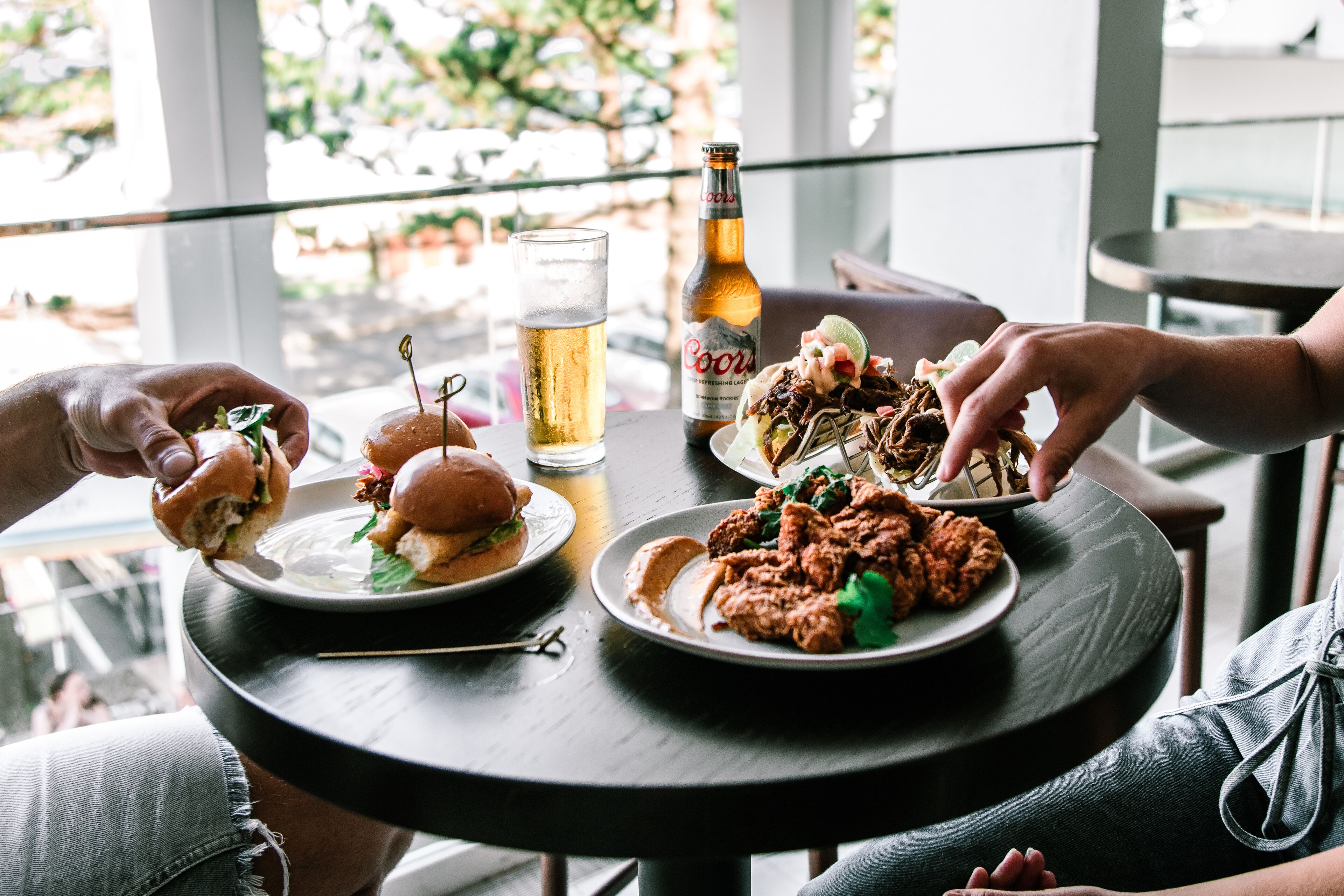 A round table with a beer bottle, a glass of beer, and plates of food including burgers and fried chicken.