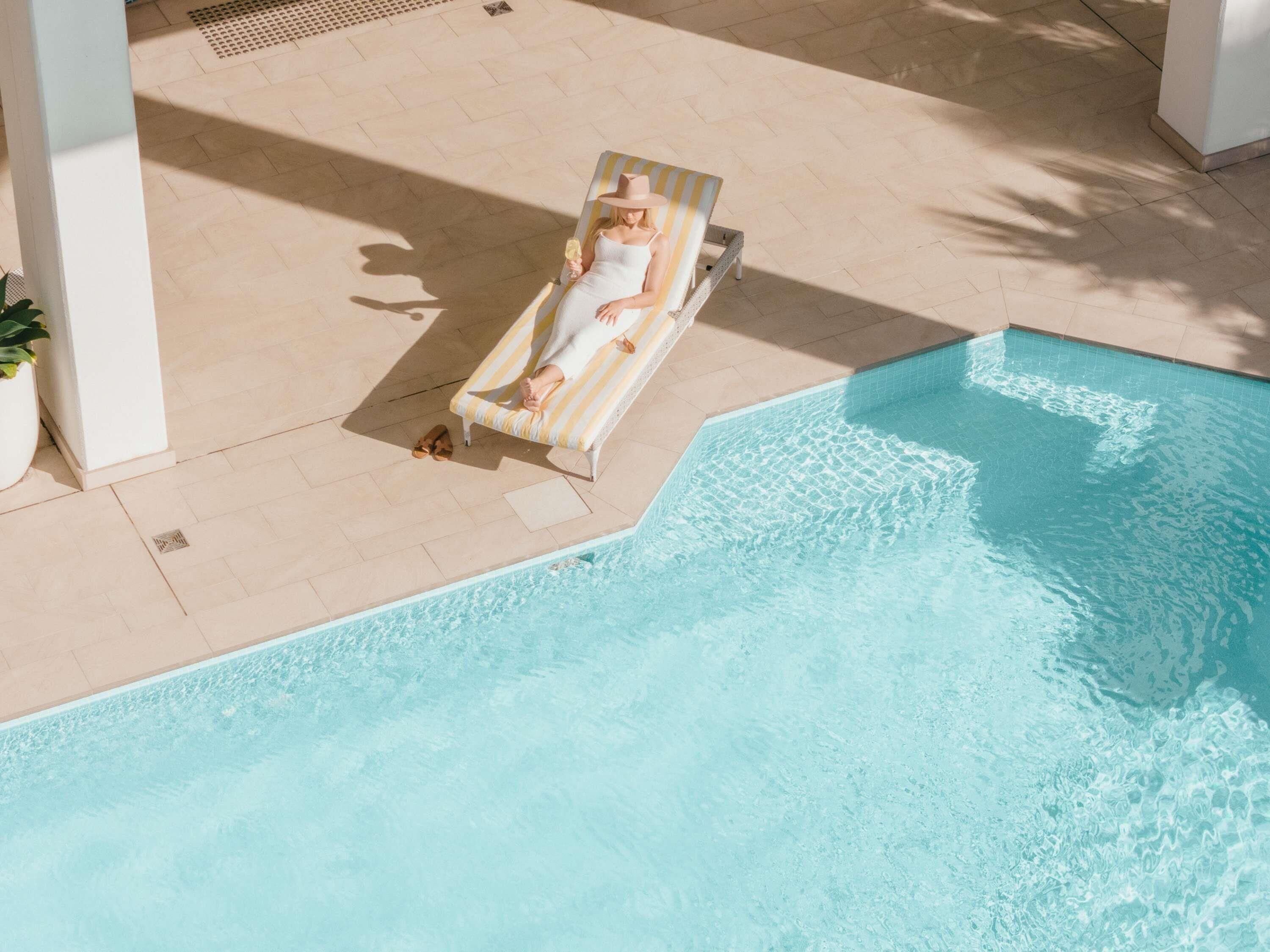 A person relaxing on a striped lounge chair by a pool with a tiled floor and a white column nearby.