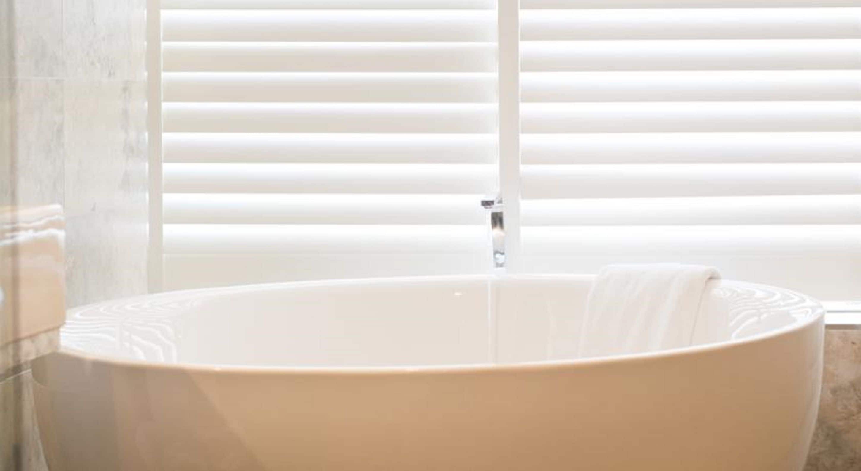 Une grande baignoire sur pieds, placée dans une salle de bain aux carreaux blancs et dotée d’une fenêtre avec des stores horizontaux.