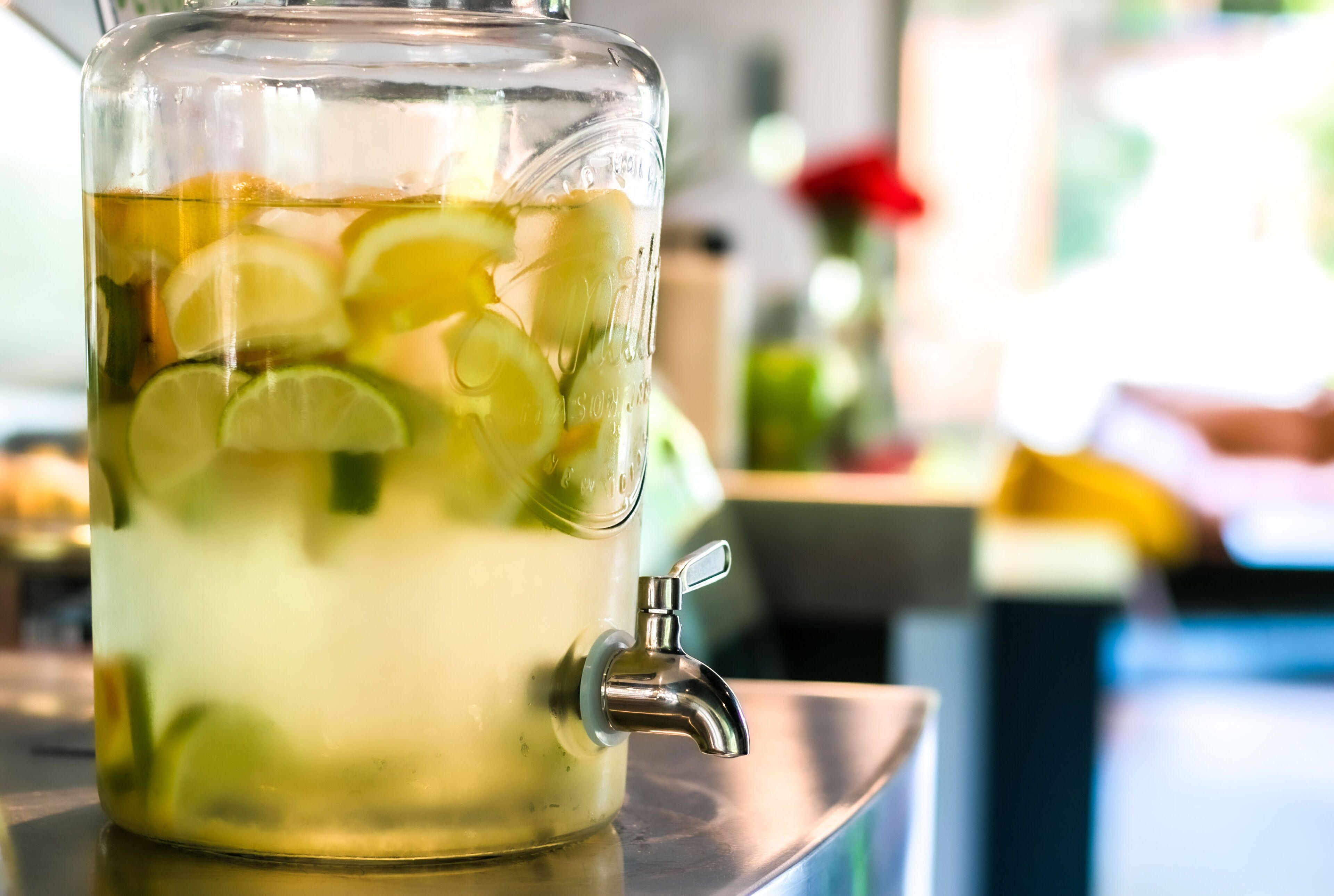 A clear glass pitcher with a tap, filled with a beverage and lime slices.
