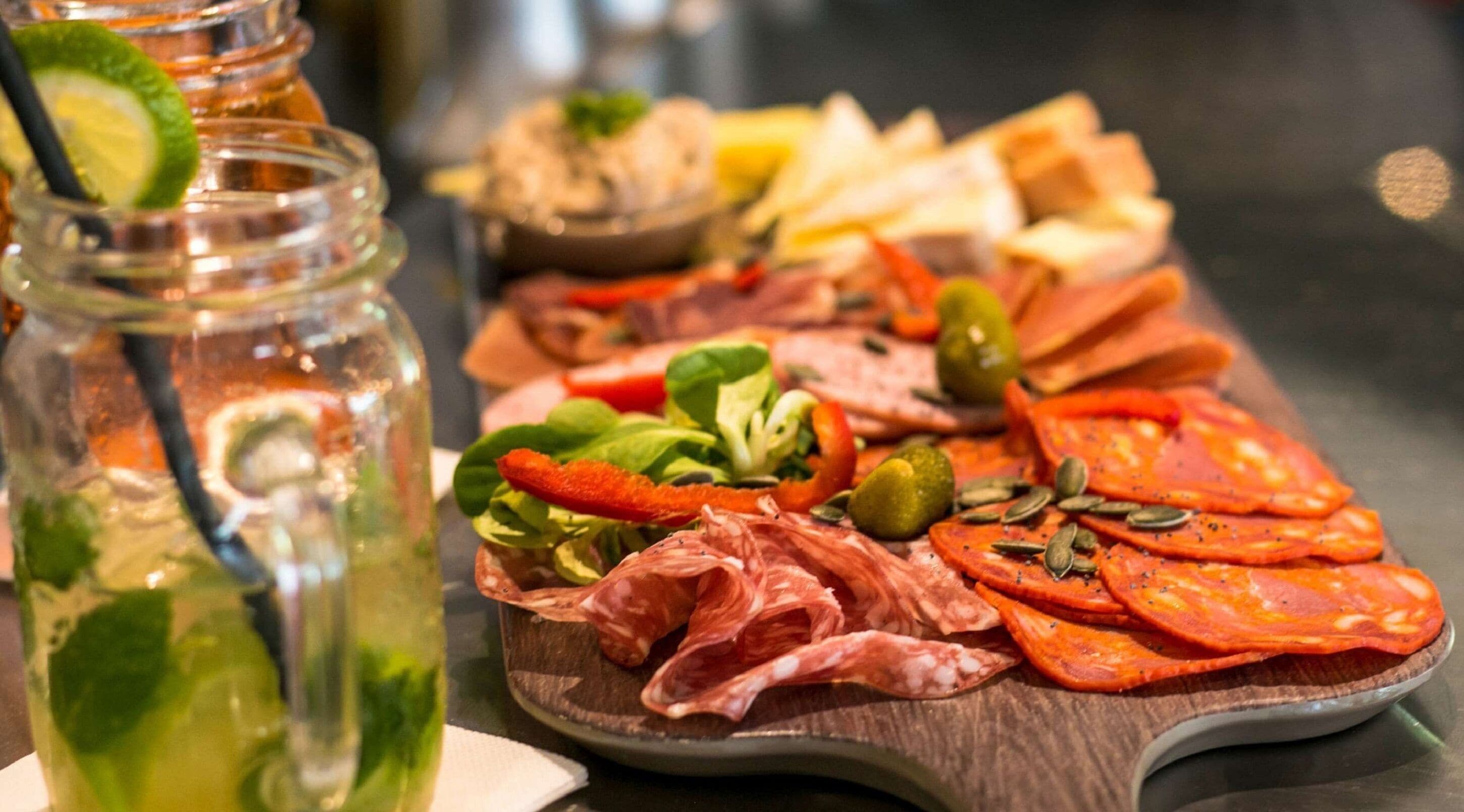 A charcuterie board with various meats, vegetables, and a jar of pickled vegetables.