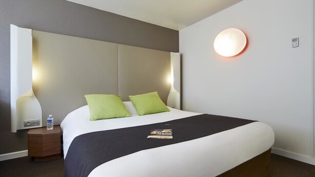 Habitación de hotel con una cama grande, dos almohadas verdes, una manta negra, una mesita de noche con una botella de agua y una lámpara fijada a la pared.