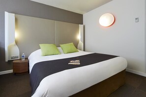 Hotelkamer met een groot bed, twee groene kussens, een zwarte plaid, een nachtkastje met een waterfles en een wandlamp.