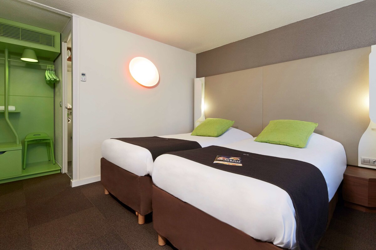 A hotel room with two beds, a green bathroom, and a white wall with a round light fixture.